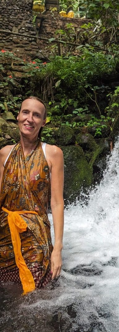 Frau in buntem Kleid steht im Wasserfall, umgeben von grüner Natur in Bali. Entspannung und Erfahrungen beim Reisen