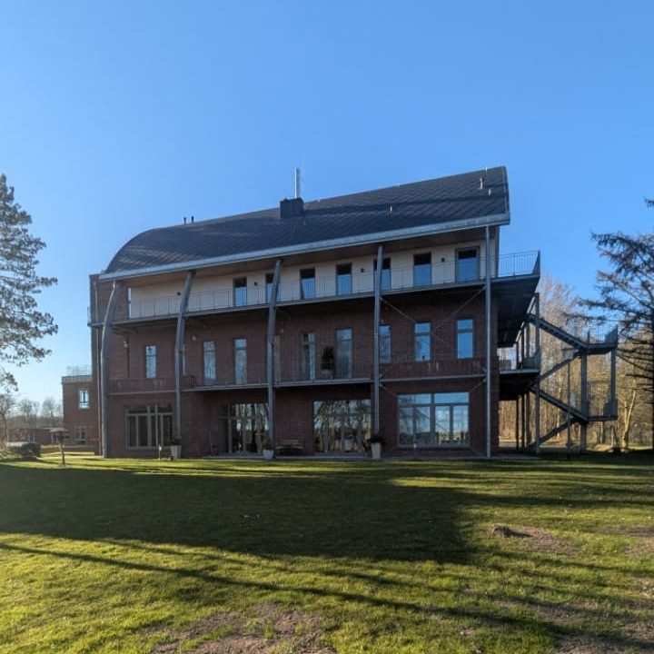 Modernes, dreigeschossiges Gebäude mit großen Fenstern und grünem Umfeld.